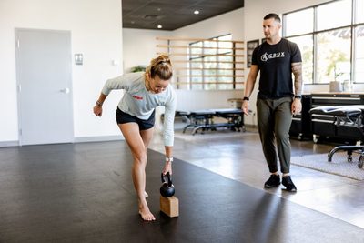 Client standing on one leg with a kettlebell in one hand leaning forward while the chiropractor watches and gives guidance